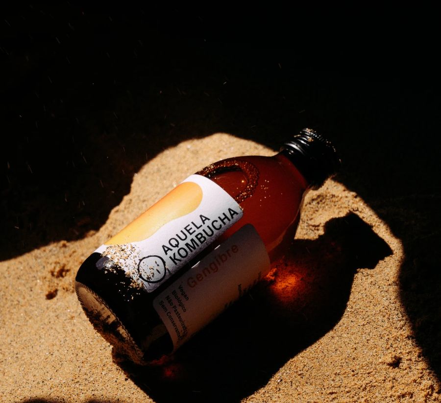 Garrafa Aquela Kombucha Gengibre deitada na areia ao sol